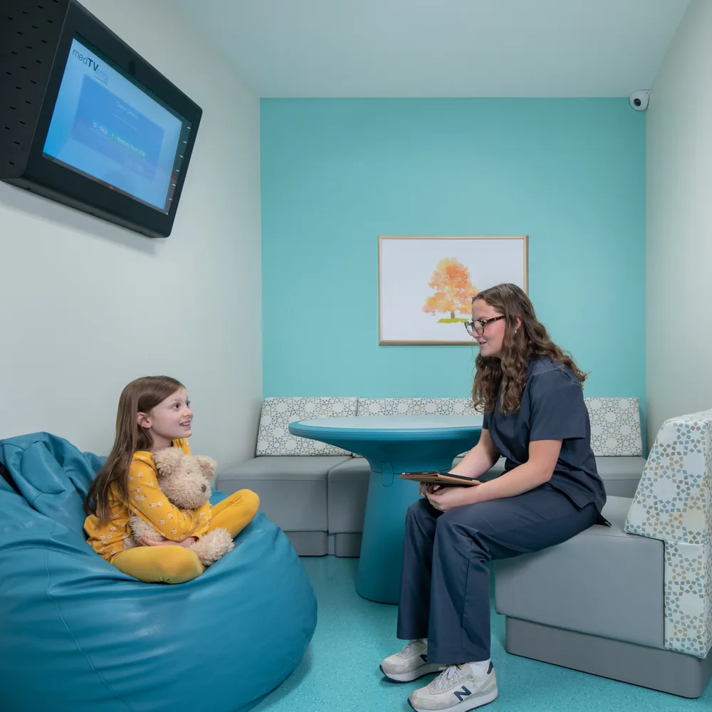A child sits with a soft toy in a large beanbag while a clinician talks with her from a bench seat, in a small assessment room finished in turquoise with a framed tree print.