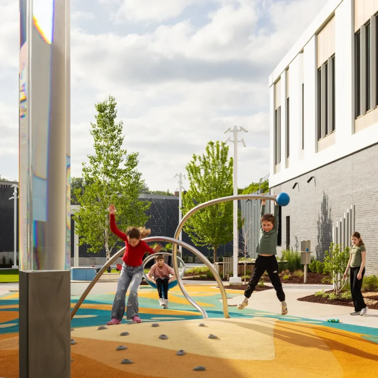 Children run and play on a coloured rubber play surface in a hospital courtyard, beside stainless steel climbing arches and a mirrored column.