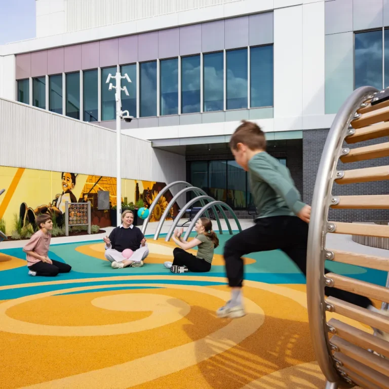 Children play in a hospital courtyard on a patterned orange and teal surface, with climbing hoops, a spinning seat and a painted mountain mural along the boundary wall.