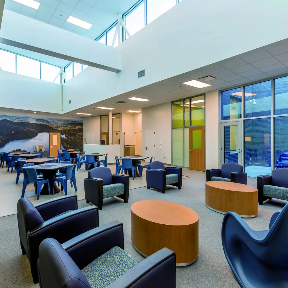 A double-height day room lit by clerestory windows, with a painted lakeside landscape mural along the far wall, dining tables beyond and a foreground cluster of blue and teal armchairs around a round table.