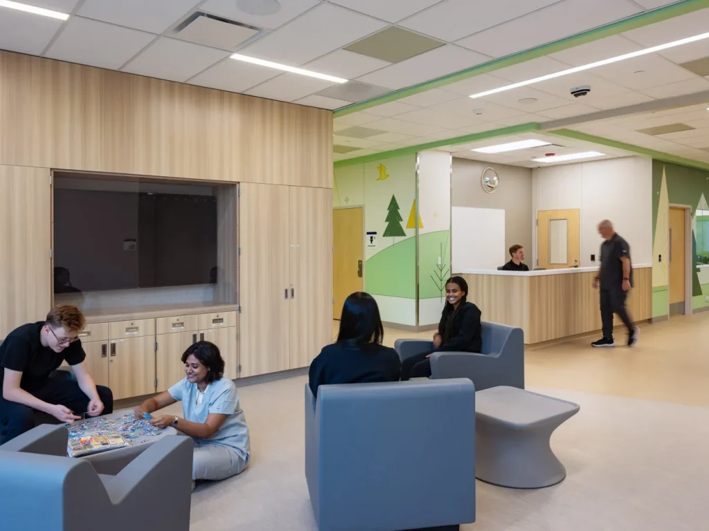 Young people and staff sit together over a puzzle in a day room finished in pale wood and green, with an open nurse station and painted forest graphics beyond.